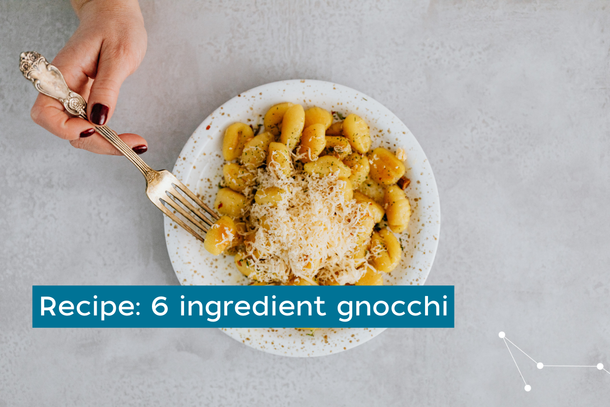 Grey background with a white and black speckled bowl filled with gnocchi and parmesan. A females hand is using a fork to pick one up