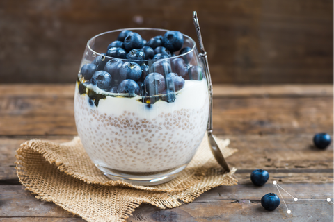 Glass cup containing chia seed mixture with blueberries on top, sitting on a beige muslin cloth on a rustic wooden table