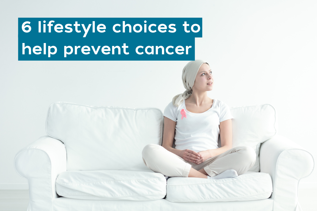 Woman dressed in white with a white scarf on head sitting on a white two seater couch with a pink breast cancer ribbon on her lefthand side chest
